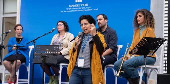 Yael Hess speaking in front of four seated musicians